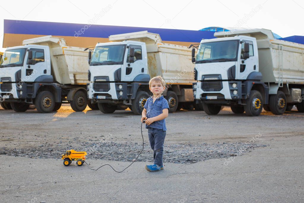 cute boy plays with a toy dump truck on control panel on asphalt. behind him are large real dump trucks. sunny warm day. Favorite games for boys