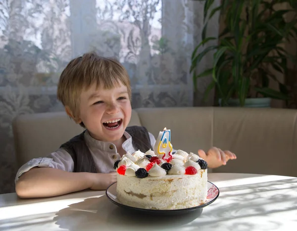 明るい誕生日の男の子は4番の形のキャンドルでクリームケーキの近くのテーブルに座っています 彼は笑いに満ちている 火災を爆破するつもりです お祭り気分 願い事をします ケーキに集中しろ — ストック写真