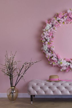 Pembe bir duvarın üzerinde çiçek açan bahar dallarının yarım çerçevesi, altında hafif bir bank, üzerinde pembe kurdeleli bir hasır şapka var. İç kısımda bahar havası, merhaba bahar, sıcak atmosfer.