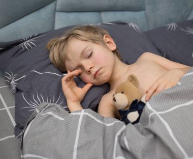 Gri bir yatakta uyuyan 4-5 yaşlarında tatlı bir çocuk. Onun yanında bir oyuncak ayı var. Tatlı sağlıklı bebek gündüz uykusu, çocuk bakımı. Günaydın, pozitif atmosfer, rahatlık ve rahatlık