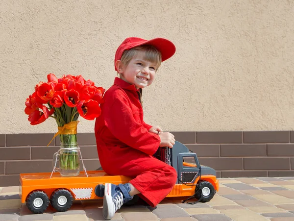 funny cute 4 years old boy in a red mechanic overalls and a cap sitting on a toy truck with large bouquet of tulips. Delivery concept, little courier, gift for women, with love for mommy. Copy space