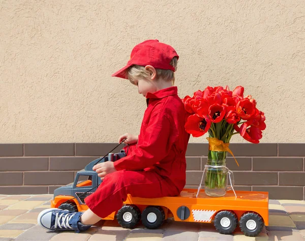 toddler boy in a red mechanic overalls and a cap sitting on a toy truck with a large bouquet of tulips. Delivery concept, little courier, a gift for women, with love for mommy