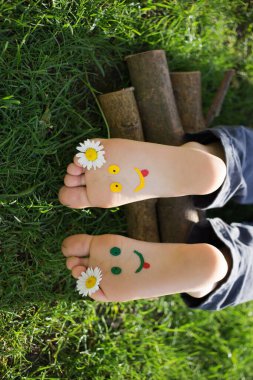 Sarı ve yeşil yüzler, çimlerin üzerinde yatan bir çocuğun çıplak ayaklarına boyanır. Papatya çiçekleri, güneşli bir gün, neşeli pozitif bir atmosfer, mutlu bir çocukluk. Merhaba yaz. çocuk sanatı