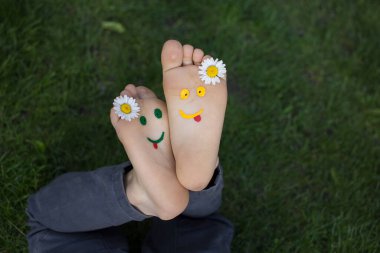 Sarı ve yeşil yüzler çimlerin üzerinde uzanan bir çocuğun çıplak ayaklarına boyanır. Papatya çiçekleri, yeşil çimenler, neşe, neşeli pozitif atmosfer, mutlu çocukluk. Merhaba yaz, gülümse gün