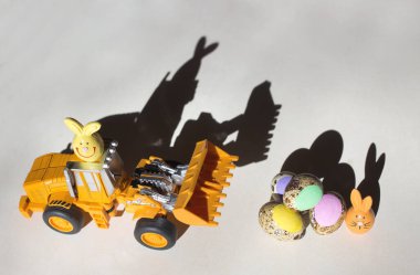 Hafif arka planda oyuncak kazıcı yükleyicisi, yanında çok renkli Paskalya bıldırcın yumurtaları ve küçük hatıra tavşanları bulunur. Karşıt gölge. İş dünyası için Paskalya tatili konsepti, inşaat şirketleri