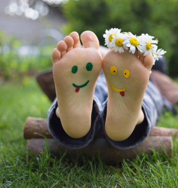 Güneşli bir günde yeşil çimlerin üzerinde uzanan bir çocuğun çıplak ayaklarına gülen yüzler çizilir. Parmaklarında papatya çiçekleri, neşeli pozitif atmosfer, mutlu çocukluk. Merhaba yaz, gülümse gün