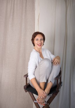 Başarılı stil sahibi, neşeli, kendine güveni olan, 30 yaşını geçmiş, hafif elbiseli bir kadın sandalyeye oturmuş, samimi bir şekilde gülümsüyor. İyi olmak, her günün neşesi, olumlu düşünmek.