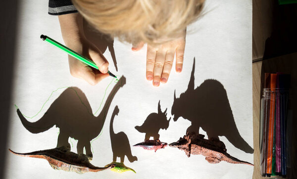 ребенок, с энтузиазмом и концентрацией, очерчивает тени игрушечной фигурки маленьких и больших динозавров. развитие мелкой моторики. развлечения для детей. Идеи для творчества для детей