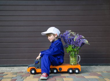 4-5 yaşlarında mavi tulumlu ve kepli bir çocuk, elinde büyük bir buket çiçekle oyuncak bir kamyonda profilde oturuyor. Teslimat konsepti, küçük kurye, kadınlara hediye, anneye sevgilerle.