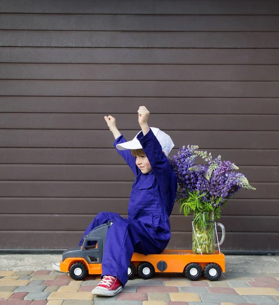 toddler boy in purple worker jumpsuit and cap sits on toy truck with bouquet of flowers , raising his hands up. Delivery concept, congratulations for mom, little courier, successful person, idea