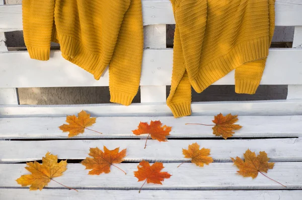 On the back of a white wooden bench hang two mustard-colored knitted sweaters. orange-red maple leaves lie nearby. Autumn atmosphere