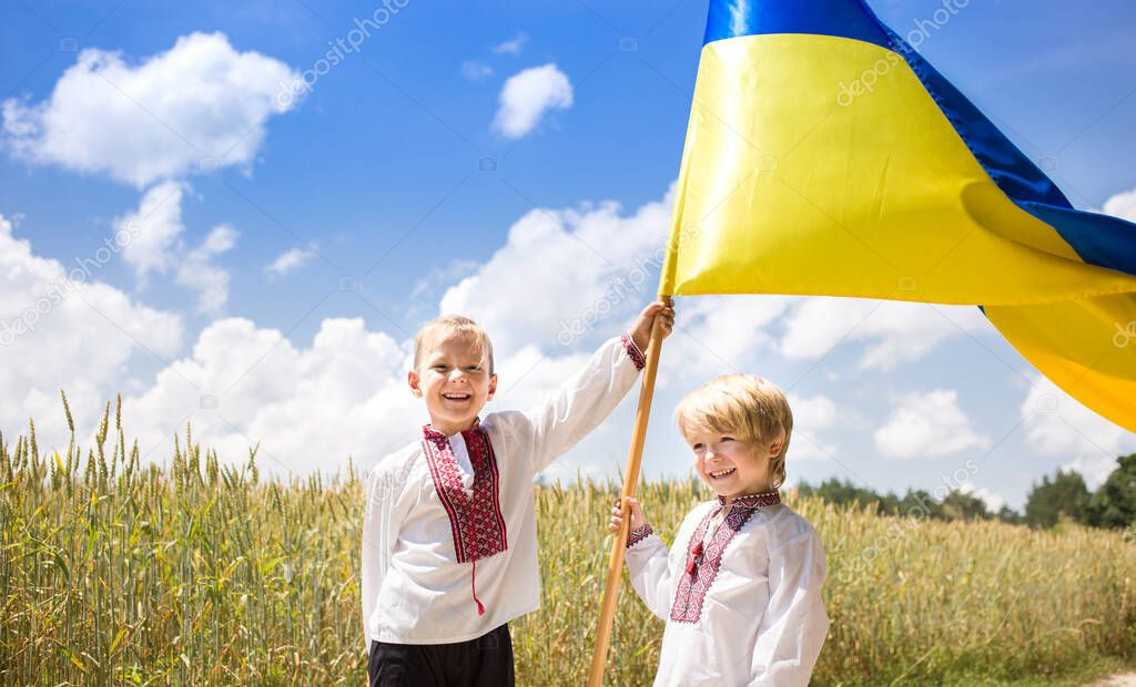 Two happy smiling boys in national Ukrainian shirts are holding a large yellow - blue flag against the background of a wheat field. Ukrainian nationality. Patriotic education. Independence Day
