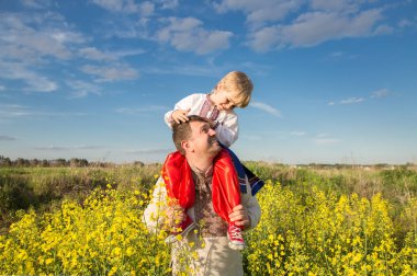 Oğlu babasının omuzlarında oturuyor. Her ikisi de ulusal Ukrayna işlemeli gömleklerini çiçek açan bir ırz tohumu tarlasının arka planına karşı giyerler. Vatansever eğitim. Gurur, özgürlük, milliyet Ukrayna