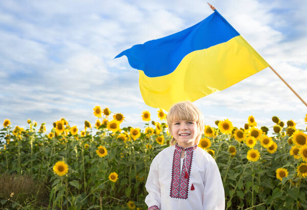 Улыбающийся мальчик в национальной украинской вышитой рубашке с желто-синим большим флагом на заднем плане. цветущее поле подсолнухов. гордость, символ страны, гражданства Украины. Патриотическое воспитание
