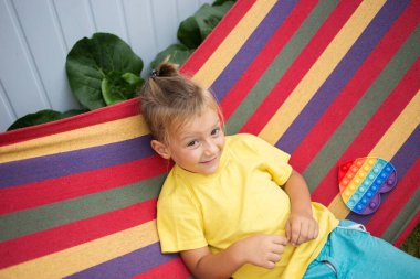boy of 5 years old sits on a multi-colored hammock, plays with a toy Pop It in the shape of a heart. The child is on vacation. trendy entertainment for children with a relaxing effect