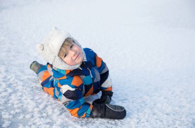 4-5 yaşlarında, kışın giydiği tulumlu sevimli bir çocuk karlı zeminde oturuyor. Çocuklarla karda aktif kış yürüyüşleri. Mevsimlik sevinçler, mutlu çocukluk. Soğuk hava..