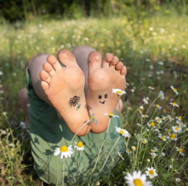Güler yüz ve çiçek, güneşli bir günde çayırda uzanan çıplak ayakla boyanır. Her yerde papatyalar açar, neşeli, pozitif bir atmosfer, mutlu bir çocukluk. Merhaba, yaz, gülümseme günü.