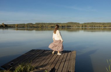 Güneşli bir günde göl kenarında açık havada dinlenmekten hoşlanan elbiseli mutlu genç bir kadın. İyilik ve olumlu bir bakış açısı. Canlılık, özgürlük ve neşe, sevgi dolu bir hayat. Doğa yürüyüşü.
