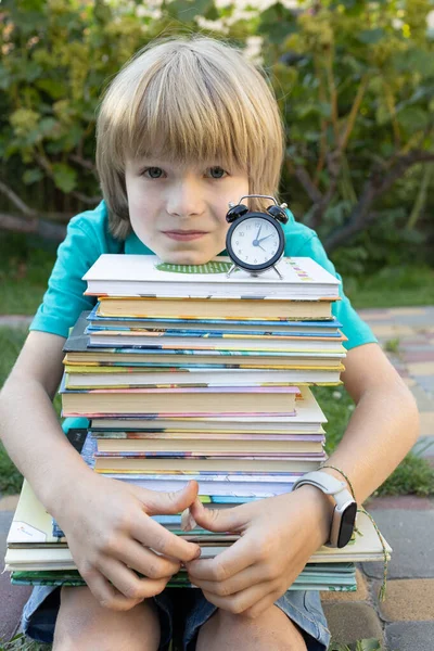 Çalışma zamanı. Küçük okul çocuğu elinde bir yığın kitap ve çalar saat tutuyor. Okuma edebiyatı, okuma hobisi. Çalışmaktan yoruldum.