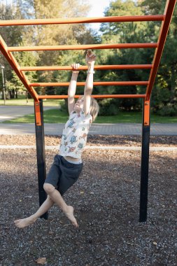Çıplak ayaklı çocuk kollarını bir yaz günü bir spor parkındaki oyun parkında yatay bir barda hareket ettiriyor. Çocuklar için eğlence ve aktiviteler. Sağlıklı çocukluk.