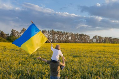 Baba, sarı kolza tohumu tarlasına karşı büyük Ukrayna bayrağı taşıyan oğlunu omuzlarında tutuyor. Vatansever eğitim. Bağımsızlık Günü. Gurur. Ukraynalılar savaşa karşı. Birlik, destek