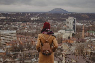Genç bir kadının kameraya karşı duruşu ve şehir manzarasına bakışı.