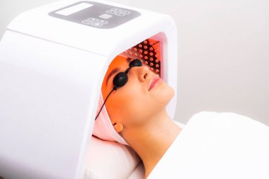 Yüze fototerapi uyguladı. Kozmetik alanında fototerapi. Takım elbiseli ve gözlüklü güzel bir kadın kozmetoloji ofisinde yüz fototerapisi prosedürü üzerine yatıyor..
