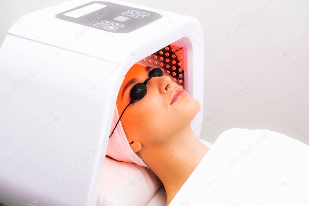 Led phototherapy for the face. Phototherapy in cosmetology. A beautiful young woman in a suit and glasses lies in the cosmetologists office on the face phototherapy procedure.