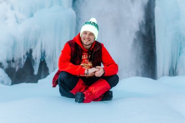 Kabardino-Balkaria, Rusya - 5 Ocak 2021: Parlak elbiseli bir gezgin kışın kar altında köpüklü bir düşüşün arka planında oturur. Şelaleye git..