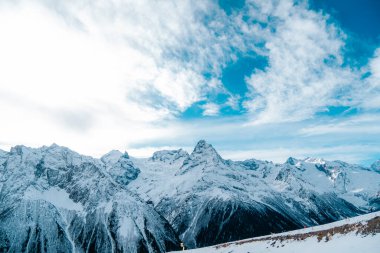 Kışın, güneşli bir havada, yakın planda dağ zirvelerinin fotoğrafı. Karlı dağlar, beyaz bulutlar, mavi gökyüzü.