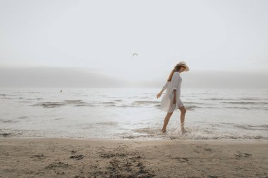 Yazın gün batımında bir kadın deniz kıyısında yürür. Plajda beyaz yazlık elbiseli bir kız..