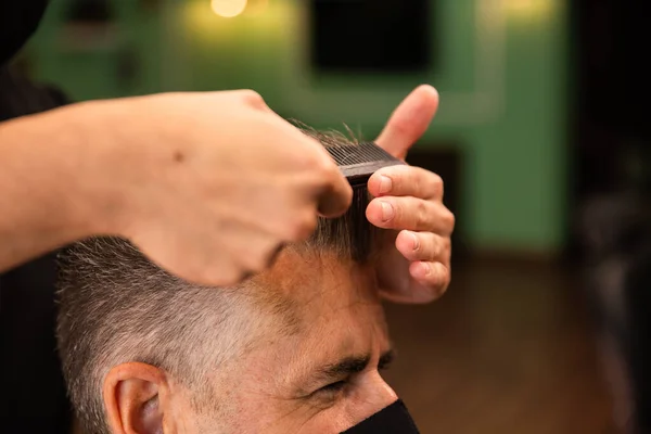 in a barber shop a barber combs a grown man with his hand and comb, they wear masks for prevention of pandemic coronavirus. the client is seated. hipster style. both are white caucasians