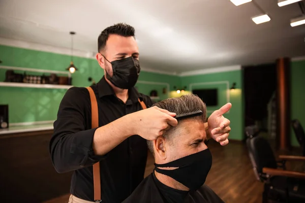 in a barber shop a barber combs a grown man with his hand and comb, they wear masks for prevention of pandemic coronavirus. the client is seated. hipster style. both are white caucasians