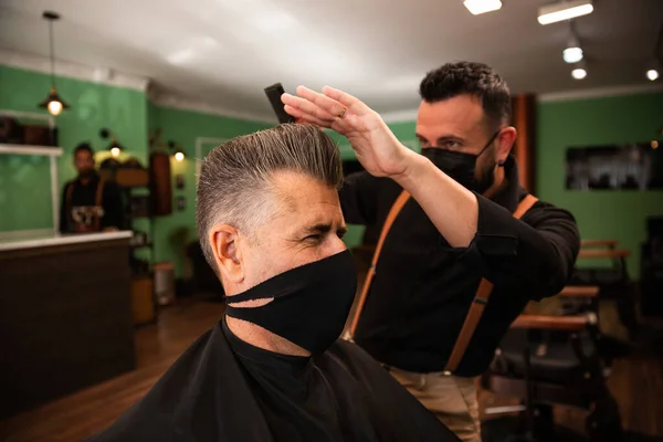 in a barber shop a barber combs a grown man with his hand and comb, they wear masks for prevention of pandemic coronavirus. the client is seated. hipster style. both are white caucasians