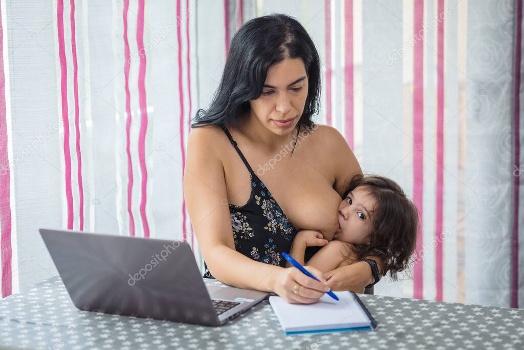 mother teleworking at the dining room table on a laptop while breastfeeding her baby girl who is sitting on her lap