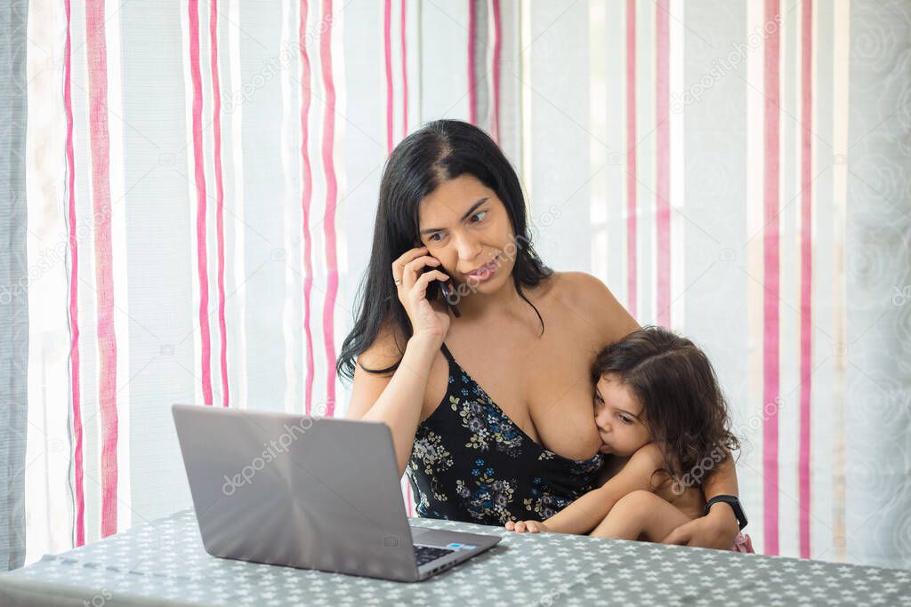 mother teleworking at the dining room table on a laptop while breastfeeding her baby girl who is sitting on her lap