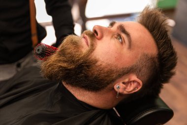 Berber dükkanında taraklı ve saç kurutma makineli İspanyol bir berber sakallı beyaz çocuğu tarıyor. Zenital düzlem.