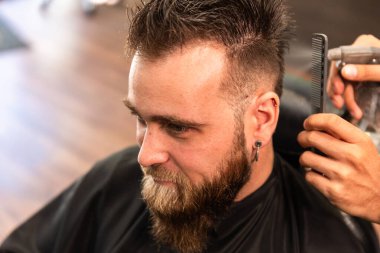 Berber dükkanında sakallı, beyaz bir adam bir Latin berberin elinden su püskürtücüsüyle taranıyor. Müşteri kulağına küpe takıyor..