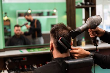 İspanyol berber, yeşil, siyah ve kahverengi berber dükkanında sakallı beyaz bir müşteriye saçlarını tarayıp kurutuyor..