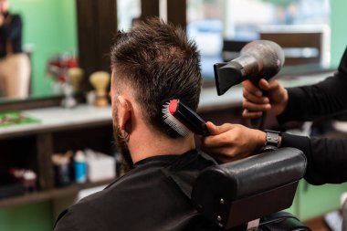 İspanyol berber, yeşil, siyah ve kahverengi berber dükkanında sakallı beyaz bir müşteriye saçlarını tarayıp kurutuyor..