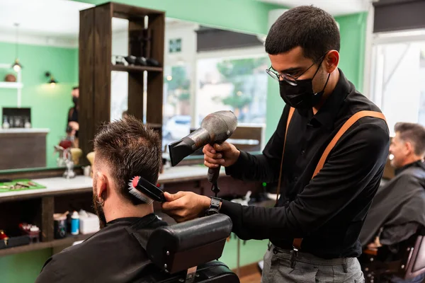 Hispanic barber combs and dries the hair of the head to Caucasian client with beard in a green, black and brown indoor barber shop.