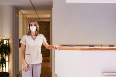 Hemşire hastanenin resepsiyonunda duruyor. Koruyucu maske takıyor.