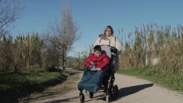 Kobieta na wózku inwalidzkim przejazd niepełnosprawne dziecko na brudnej drodze. — Wideo stockowe