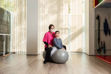 Fıstık Beden Eğitimi Balosu 'nun üstünde denge egzersizleri yapan engelli bir çocuk ve fizyoterapist. salgın maskesi koruması