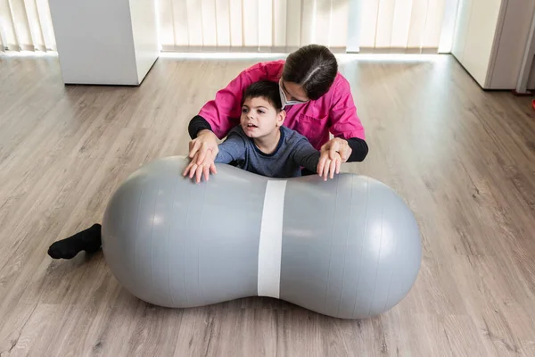 Niño discapacitado y fisioterapeuta en un Peanut Gym Ball haciendo ejercicios de equilibrio. protección máscara pandémica — Foto de Stock