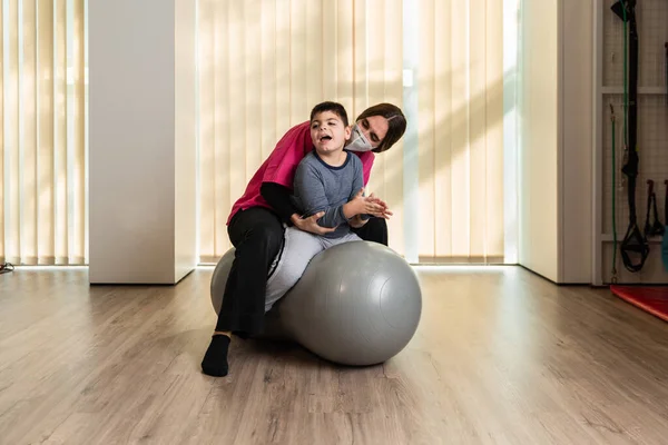 Criança deficiente e fisioterapeuta em cima de uma bola de ginástica de amendoim fazendo exercícios de equilíbrio. proteção da máscara pandémica — Fotografia de Stock