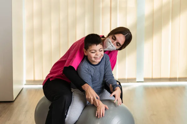 Criança deficiente e fisioterapeuta em cima de uma bola de ginástica de amendoim fazendo exercícios de equilíbrio. proteção da máscara pandémica — Fotografia de Stock