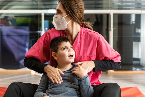 Mozgássérült gyermek és fizioterapeuta egy piros tornatermi szőnyegen gyakorlatokat végez. pandémiás maszkvédelem — Stock Fotó