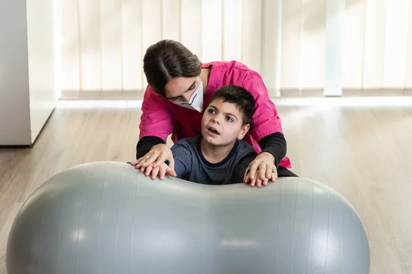 Mozgássérült gyermek és fizioterapeuta egy mogyoró tornatermi bálon egyensúlyozó gyakorlatokat végez. pandémiás maszkvédelem — Stock Fotó