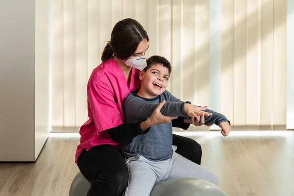 Disabled child and physiotherapist on top of a Peanut Gym Ball doing balance exercises. pandemic mask protection — Stock Photo, Image